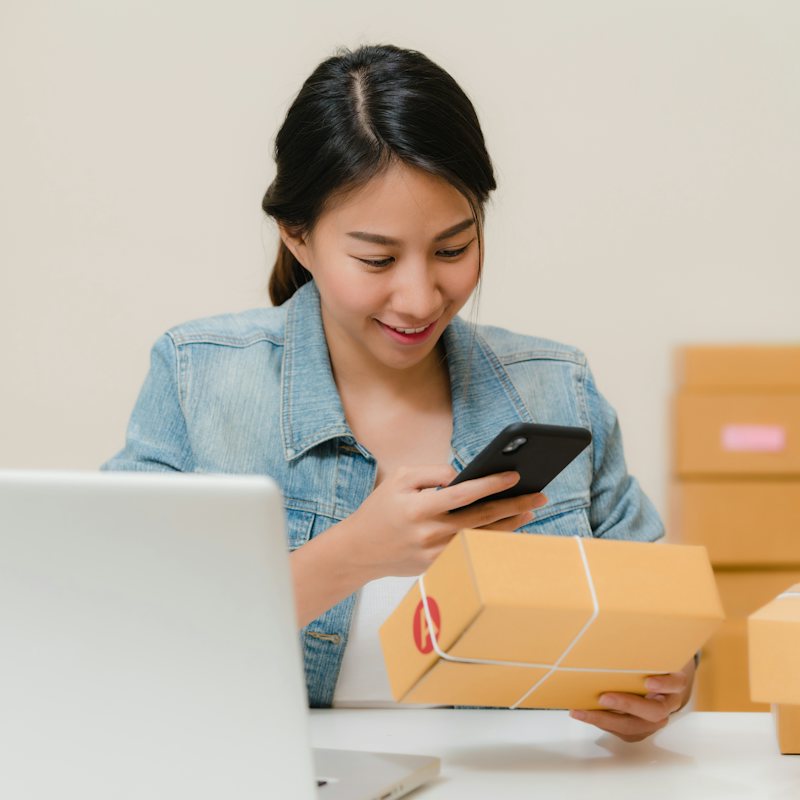 Mujer revisando un pedido en su celular junto a cajas listas para enviar