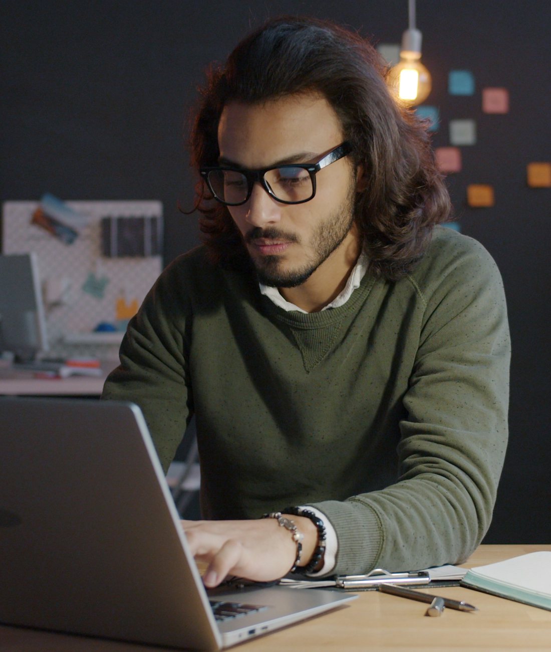 Hombre con lentes trabajando de noche en su laptop, con una libreta al lado